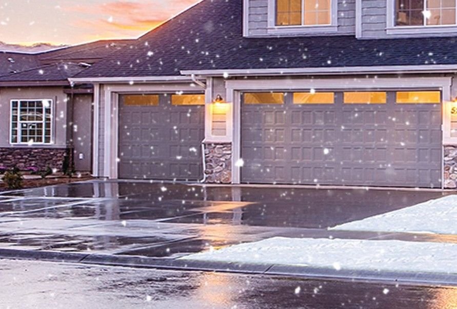 Driveway in front of a two-car garage at dusk while snow falls, the pavement wet and clear with snow only along the edge