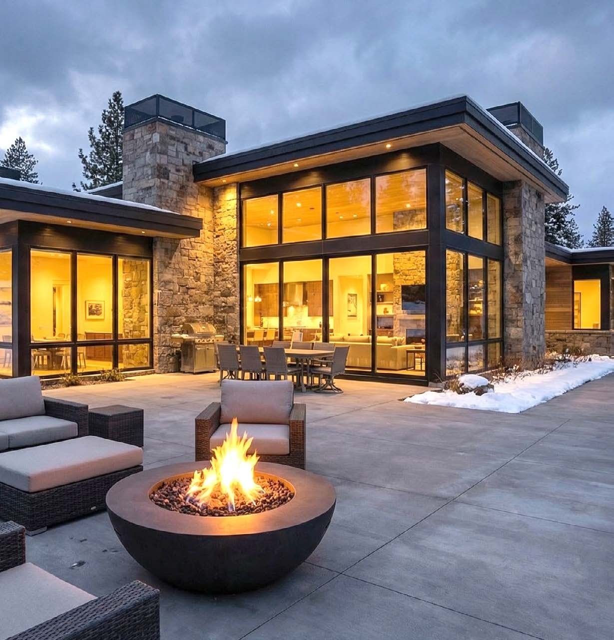 Modern stone and glass home at dusk in winter, warmly lit windows, snow on the roof and along the edge of a clear concrete patio